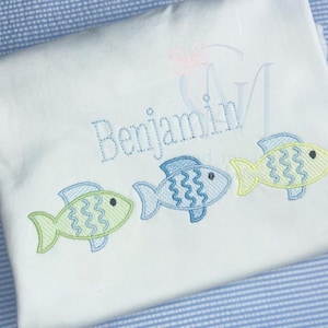 May include: White baby onesie with the name "Benjamin" embroidered in blue thread. Three embroidered fish are in a row below the name, one blue, one green, and one yellow.