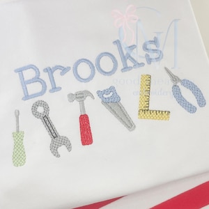 Könnte beinhalten: Weißer Stoff, bestickt mit dem Namen "Brooks" in Hellblau, zusammen mit Werkzeugillustrationen. Die Werkzeuge umfassen einen Schraubendreher, einen Schraubenschlüssel, einen Hammer, eine Säge, ein Lineal und eine Zange, alle in passenden Farben.
