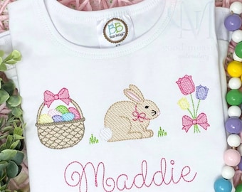 May include: White baby onesie with pink embroidered Easter bunny, basket of eggs, and flowers. The onesie is personalized with the name "Maddie".