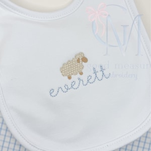 May include: White baby bib with a light brown embroidered sheep and the name "Everett" in blue stitching. The bib has a blue and white checkered pattern on the bottom.