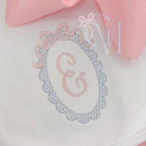 May include: White fabric with a pink embroidered letter "E" inside a blue scalloped oval frame, adorned with pink bows. A large pink bow is visible in the background. The design is delicate and feminine.