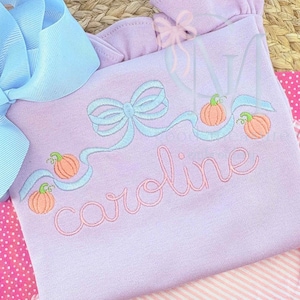 Op de afbeelding: Een lichtpaarse babyoutfit met een lichtblauw strik- en lintontwerp, met geborduurde pompoenen. De naam "Caroline" is in een cursief lettertype geborduurd. De outfit heeft een gerimpelde halslijn en staat op een roze en wit gestreepte achtergrond.
