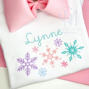 Op de afbeelding: Wit shirt met de naam "Lynne" geborduurd in turquoise draad. Het shirt is versierd met meerdere geborduurde sneeuwvlokken in verschillende kleuren, waaronder paars, roze, turquoise en blauw.