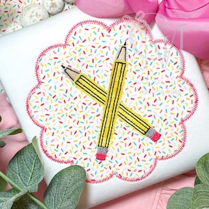 May include: A white fabric with a pink scalloped border featuring two yellow pencils crossed over each other. The background is a white fabric with a pattern of colorful sprinkles.