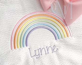 Może przedstawiać: Biały haftowany kocyk dla dziecka z wzorem tęczy i imieniem "Lynne" wyszytym fioletową nicią.