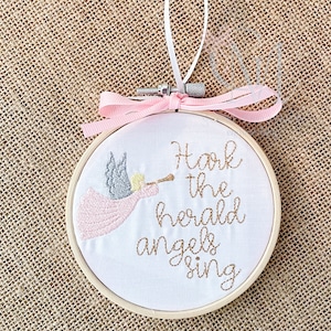 May include: A white embroidery hoop with a pink and silver embroidered angel playing a trumpet. The text "Hark the herald angels sing" is embroidered in gold thread.