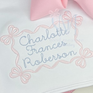 Op de afbeelding: Witte stof met geborduurde naam "Charlotte Frances Roberson" in blauw, omlijst door een roze lint en strikken. Een grote roze strik is zichtbaar op de achtergrond. Delicaat en vrouwelijk ontwerp.