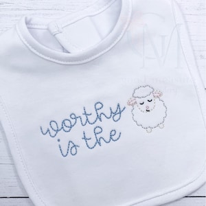 May include: White baby bib with a blue embroidered text that reads "worthy is the" and a cartoon sheep illustration. The bib has a rounded neckline and a snap closure at the back.