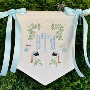 May include: A white banner with blue ribbon ties, featuring embroidered storks carrying bundles, a wreath of green leaves, and the monogram "BTM".