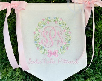 May include: A white banner with pink ribbon ties. The banner features a pink monogram "SPW" surrounded by a wreath of green leaves and colorful flowers. The text "Sadie Nelle Pittard" is embroidered below the monogram.