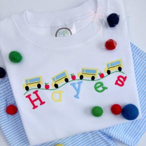 May include: A white short-sleeved t-shirt featuring an embroidered design of three yellow school buses and red apples on a green wavy line. Below the buses, the name Hayes is embroidered in large, multi-colored letters: red, yellow, blue, green, and red.