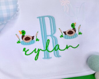 May include: A white bib with a blue and green embroidered letter R, the name "rylan" in green cursive, and two embroidered ducks with green heads and brown bodies.