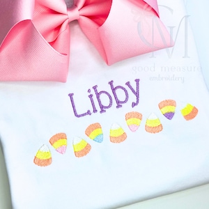Może przedstawiać: Biała koszulka z wyhaftowanym imieniem "Libby" fioletową nicią. Poniżej imienia znajdują się haftowane wzory cukierków kukurydzianych w kolorze pomarańczowym, żółtym i białym. Duża różowa kokarda znajduje się w lewym górnym rogu.