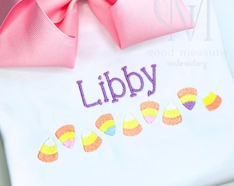 May include: White shirt embroidered with the name "Libby" in purple thread. Below the name are embroidered candy corn designs in orange, yellow, and white. A large pink bow is in the upper left corner.
