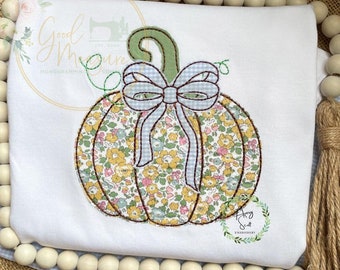 May include: White long-sleeved shirt featuring an embroidered pumpkin design. The pumpkin is adorned with a floral pattern in yellow, pink, and green, a blue gingham bow, and a green stem. The design is outlined with brown stitching.