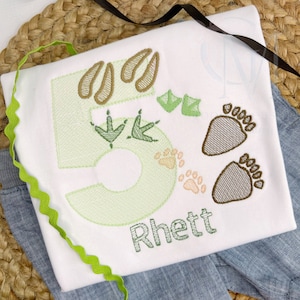 May include: A white onesie with green and brown embroidery of the number 5, animal footprints, and the name "Rhett".