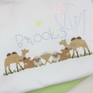 May include: White fabric with an embroidered design featuring camels, sheep, and a star. The name "Brooks" is embroidered in light blue. The design has a religious theme, with a manger scene.