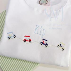 May include: White long-sleeved shirt with embroidered golf carts in blue, red, navy, and yellow. The name "Kip" is embroidered in light blue. The shirt has a round neckline and a small tag at the top.