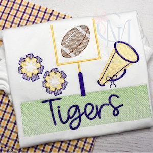May include: A white embroidered shirt with a football, goal post, megaphone, and two purple and yellow flowers. The text "Tigers" is embroidered in purple on a green field.