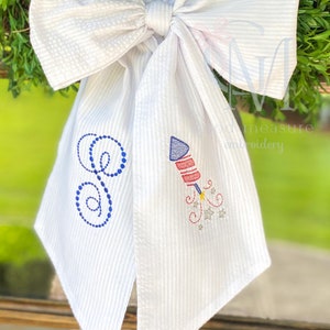 May include: White bow with blue and red embroidered letter G and a firework design. The bow is made of a white fabric with a striped pattern. The text "Good Measure Embroidery" is visible in the background.