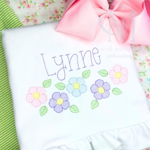 May include: White embroidered baby onesie with the name "Lynne" stitched in purple thread. The onesie has a row of five embroidered flowers in pink, purple, blue, and yellow with green leaves.