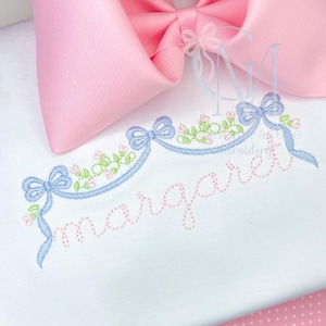Può includere: Un tessuto bianco con un fiocco rosa e dettagli ricamati. Il ricamo include un motivo a nastro azzurro con fiori rosa e il nome "margaret" in corsivo rosa.