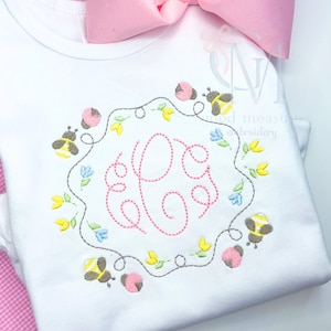 Puede incluir: Una camiseta blanca con un monograma rosa rodeado de una corona de flores, abejas y mariquitas bordadas. El texto "good measure embroidery" es visible en el fondo.
