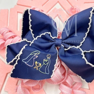 May include: A large navy blue hair bow with white scalloped edges. The bow features an embroidered nativity scene and the text "good measure embroidery". Pink and white ribbon curls are visible in the background.