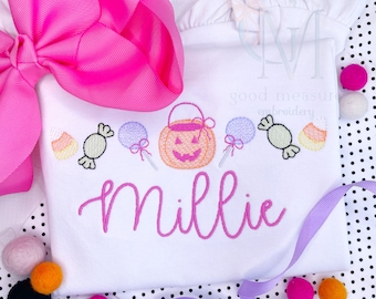 May include: White onesie with pink embroidery of a pumpkin, candy, and lollipops. The onesie is personalized with the name "Millie" in pink.