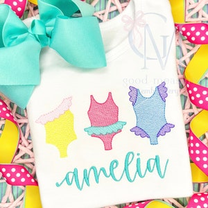 May include: A white onesie with three embroidered swimsuit designs in yellow, pink, and blue. The name "amelia" is embroidered in turquoise below the swimsuits.