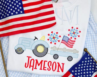 May include: White t-shirt featuring a light blue tractor design pulling a wagon with fireworks and an American flag. The name "JAMESON" is embroidered in red. Two American flags are placed on either side of the shirt.