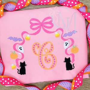 Könnte beinhalten: Rosa Halloween-Shirt mit einem großen, orange-gelben Buchstaben "C" in der Mitte. Das Shirt zeigt gestickte Halloween-Elemente, darunter eine Schleife, Fledermäuse, Maisbonbons, Geister, Kürbisse und schwarze Katzen. Ein rosa-orangefarbenes Polka-Dot-Band umrahmt das Shirt.