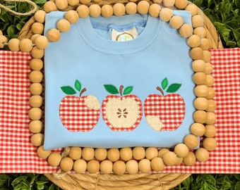 Könnte beinhalten: Ein hellblaues Sweatshirt mit drei gestickten roten und weißen Gingham-Äpfeln auf der Vorderseite. Die Äpfel haben grüne Blätter und Stiele.
