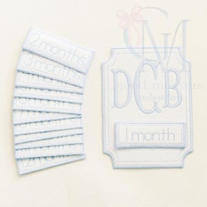 May include: White fabric baby milestone markers with light blue stitching. Includes rectangular labels with months 2-12 and a larger sign with the text "1 month" and the initials "DCB".