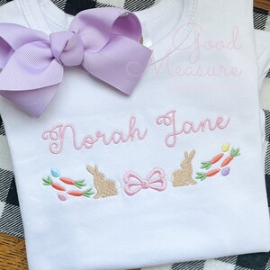 May include: White onesie with the name "Norah Jane" embroidered in pink. The onesie features a pink bow and two brown bunnies with carrots and green leaves embroidered below the name.