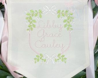 May include: A white banner with pink ribbon ties, featuring a floral wreath with a pink embroidered name "Libby Grace Cauley" in a cursive font.