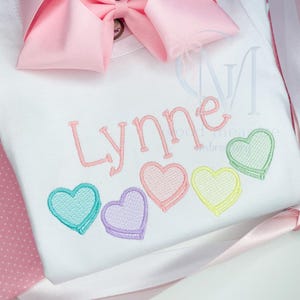 May include: A white onesie with the name "Lynne" embroidered in pink.  Five pastel-colored candy hearts are embroidered below the name.