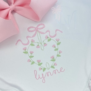 May include: White embroidered baby onesie with a pink floral heart design and the name "Lynne" stitched below.