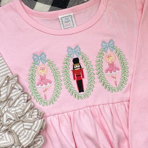 May include: Pink long-sleeved shirt with embroidered images of a Nutcracker and two ballerinas in wreaths. The shirt has a ruffled bottom and says "Sew Sweet Boutique" on the tag.