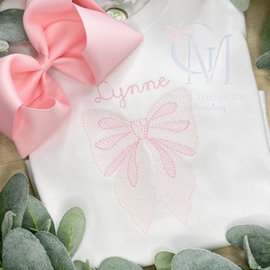 May include: White baby onesie with a pink embroidered bow and the name "Lynne" stitched in pink thread. The onesie is decorated with a pink bow and the text "Sweet measure embroidery".