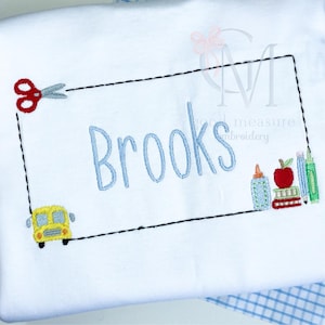 Puede incluir: Camiseta blanca con un rectángulo cosido en negro que contiene el nombre "Brooks" en azul claro. El diseño incluye unas tijeras rojas, un autobús escolar amarillo y útiles escolares.