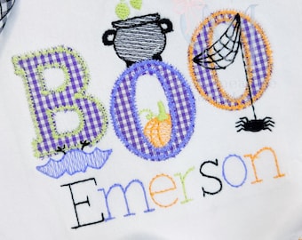 May include: White onesie with the word "BOO" in purple gingham letters, a cauldron, a spider web, and a spider. The letter "O" has a pumpkin. The name "Emerson" is embroidered below.