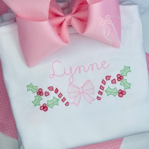 May include: White onesie with pink embroidery of a bow, candy canes, and holly leaves. The name "Lynne" is embroidered above the design.