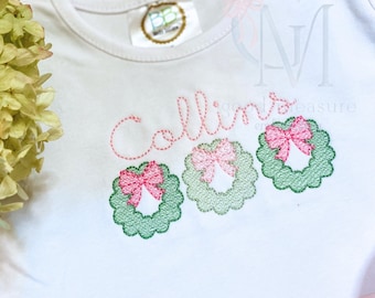 Op de afbeelding: Witte romper met de naam "Collin" geborduurd in roze draad. Drie groene kransen met roze strikken zijn onder de naam geborduurd.