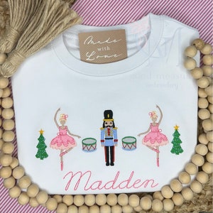 May include: White long-sleeve shirt with pink embroidery of two ballerinas, a nutcracker, and two Christmas trees. The shirt also has the text "Made with Love" and "Madden" embroidered on it.