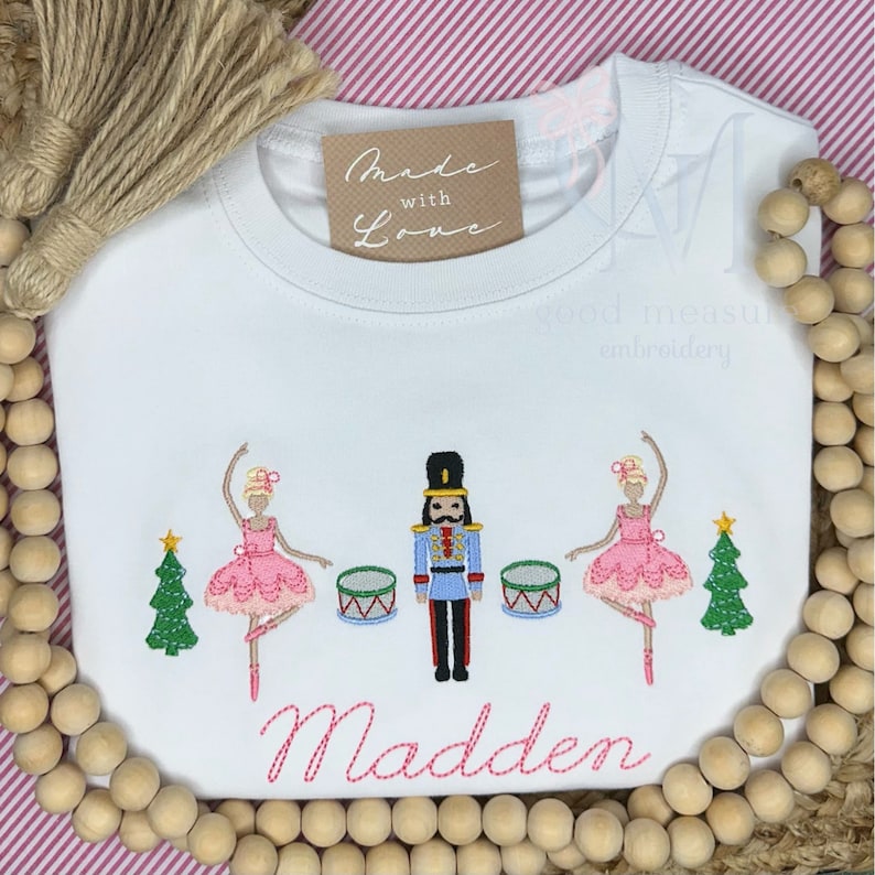 May include: White long-sleeve shirt with pink embroidery of two ballerinas, a nutcracker, and two Christmas trees. The shirt also has the text "Made with Love" and "Madden" embroidered on it.