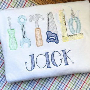 May include: A white onesie with embroidered tools and the name "Jack". The tools include a screwdriver, wrench, hammer, saw, ruler, and pliers. The tools are embroidered in various colors, including green, blue, orange, and yellow.