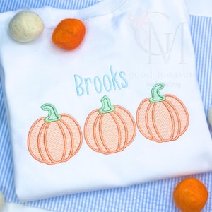 May include: White baby onesie with the name "Brooks" embroidered in blue and green thread. Three orange pumpkins are embroidered below the name.