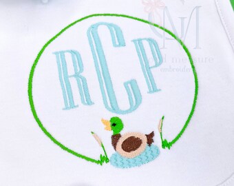 May include: A white bib with a green circle containing the monogram "RCP" in light blue and a brown duck with a blue head and yellow beak in a blue nest with green reeds.