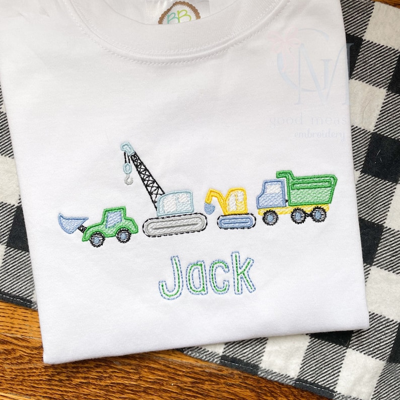 May include: A white t-shirt with an embroidered design of construction vehicles, including a crane, a backhoe, and a dump truck. The name "Jack" is embroidered below the vehicles.